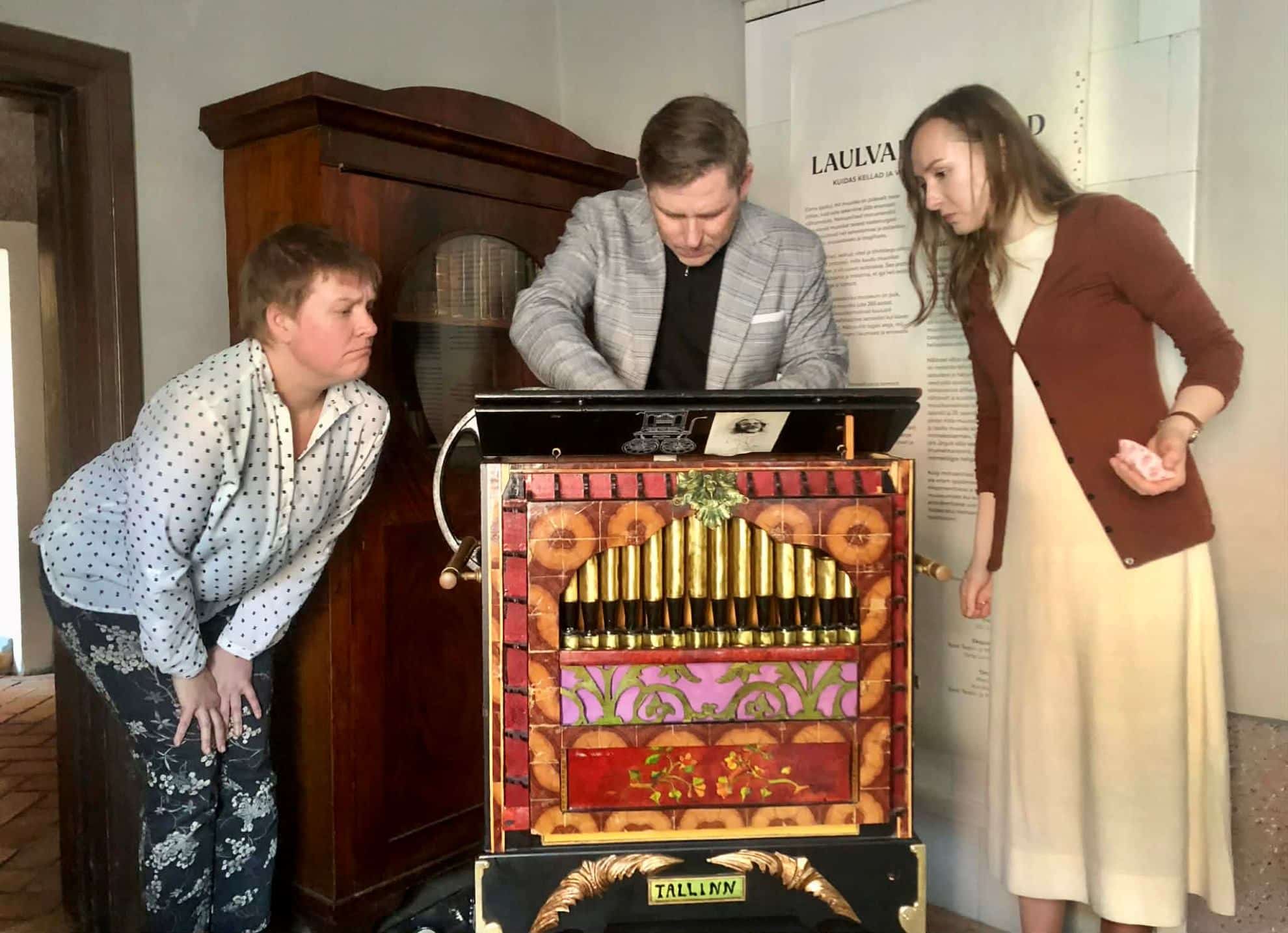 Kolm inimest uurivad linnakodaniku muuseumis vana väntmehhanismiga muusikariista; mees keskel keerab mehhanismi, kaks naist vaatavad kõrvalt