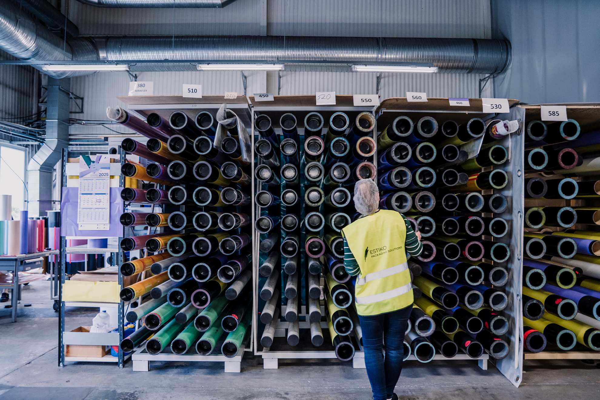 Inimene seisab tehase laos suurte värviliste materjalirullide riiulite ees ja valib nende hulgast sobivat
