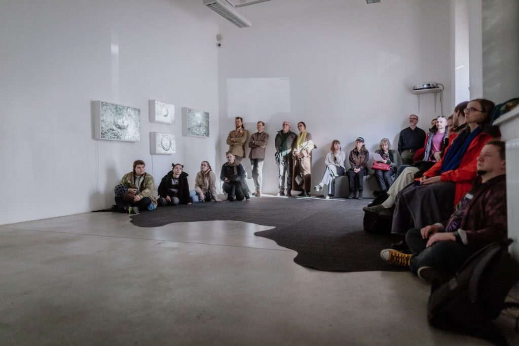 Inimesed istuvad ja seisavad Tartu Kunstimaja näitusesaalis, vaadates seintel olevaid kunstiteoseid.