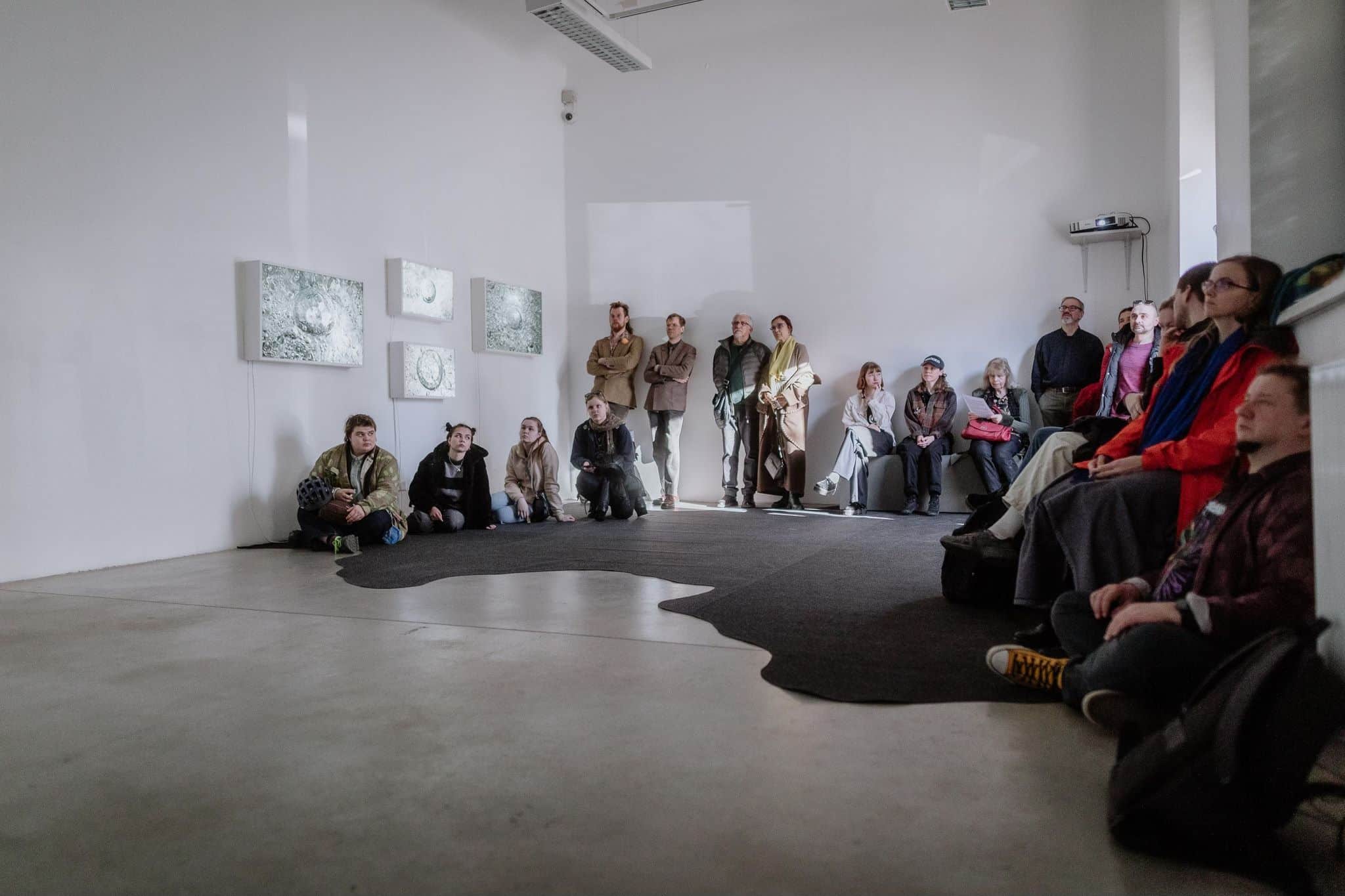 Inimesed istuvad ja seisavad Tartu Kunstimaja näitusesaalis, vaadates seintel olevaid kunstiteoseid.