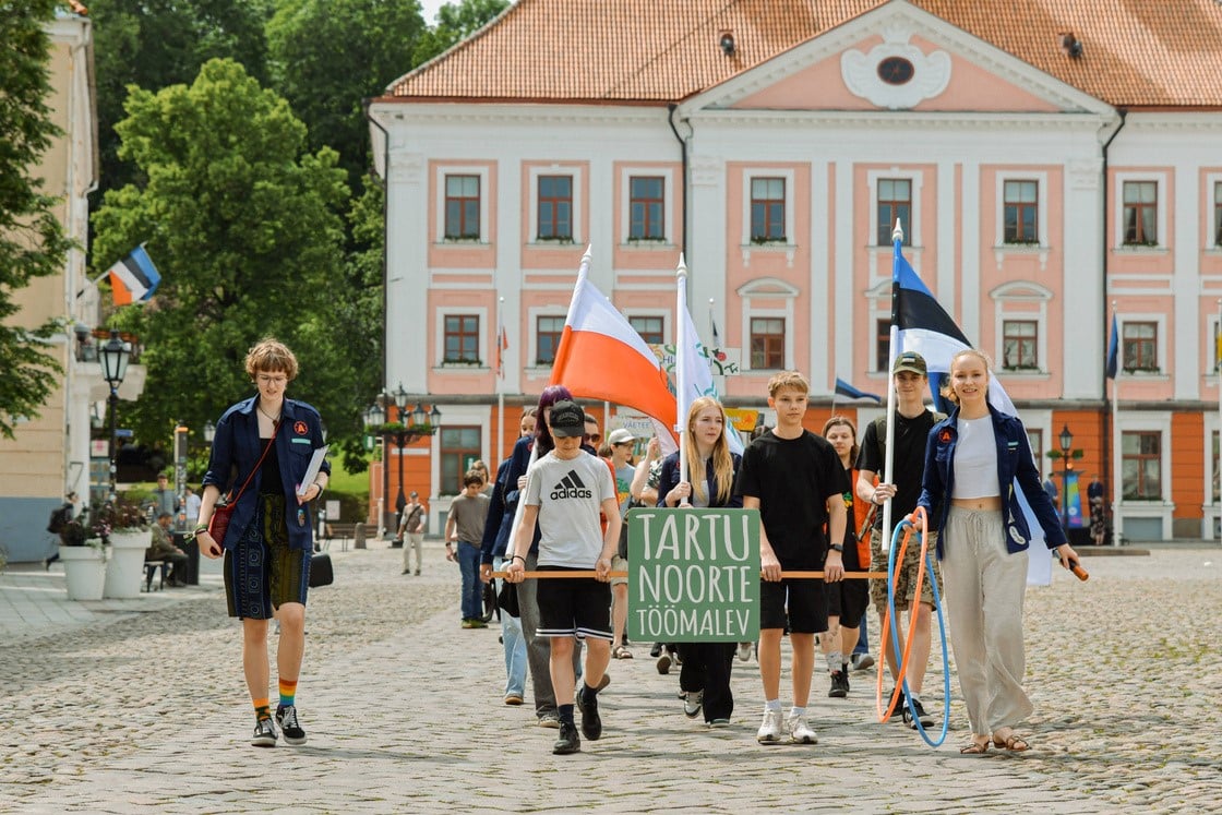 Noored marsivad Tartu Raekoja platsil, kandes silti „Tartu noorte töömalev“ ja lippe, taustal Tartu raekoda.