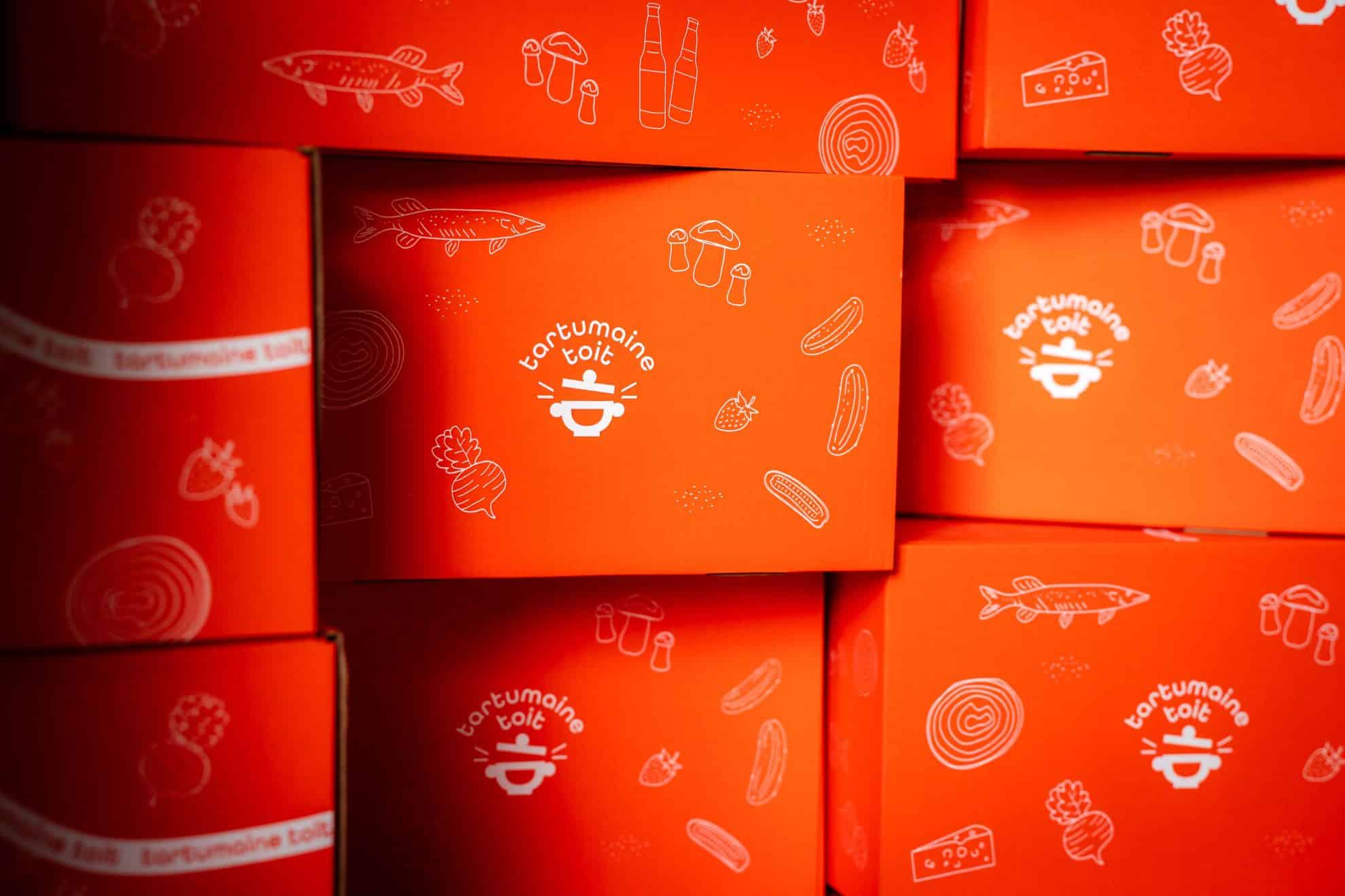 Tartumaine toit kinkekarbid oranžil taustal, karpidel illustratsioonid kalast, köögiviljadest ja kohalikust toorainest