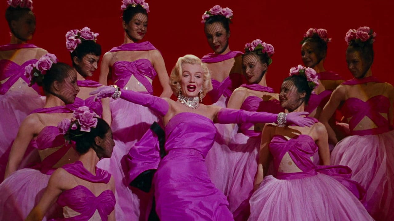 Filmikaader muusikalist, kus roosas kleidis blond laulja, Marilyn Monroe on ümbritsetud roosades kostüümides tantsijatest punasel laval.