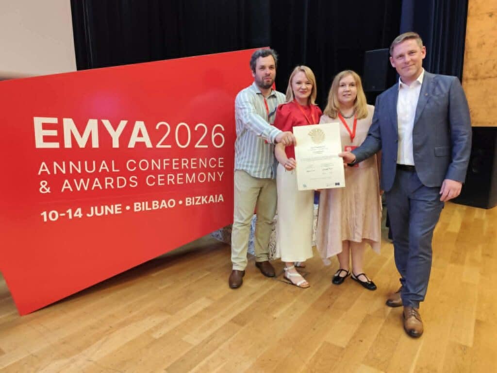 Neli inimest seisavad EMYA 2026 auhinnatseremoonia punase tausta ees ja hoiavad käes tunnustust. Tartu Linnamuuseum pälvis Euroopa aasta muuseumi auhindade jagamisel žürii eripreemia kogukonna kaasamise eest.