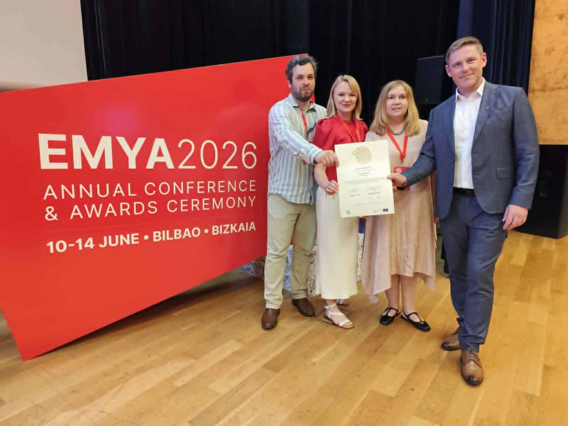 Neli inimest seisavad EMYA 2026 auhinnatseremoonia punase tausta ees ja hoiavad käes tunnustust. Tartu Linnamuuseum pälvis Euroopa aasta muuseumi auhindade jagamisel žürii eripreemia kogukonna kaasamise eest.