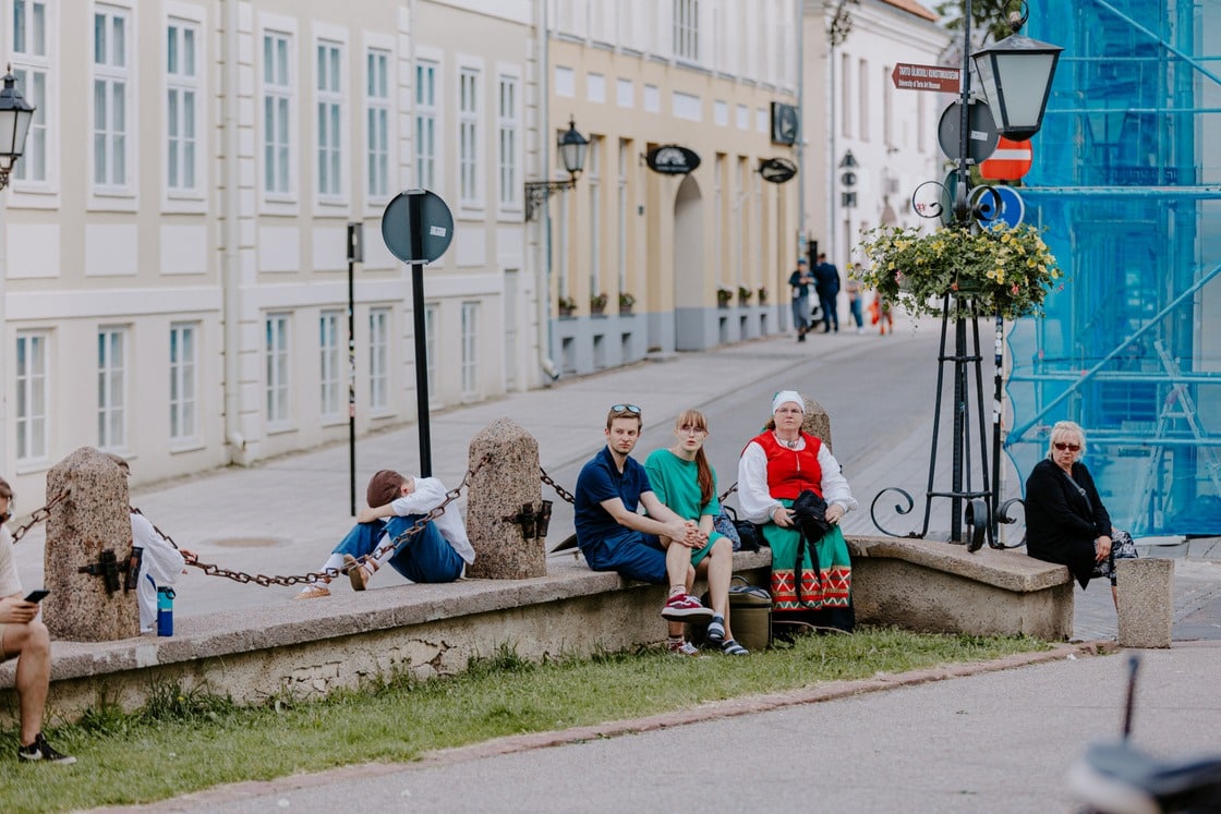 Inimesed istuvad Tartu vanalinnas Ülikooli tänava ääres madalal müüril, taustal ajaloolised hooned ja Tartu Ülikooli muuseum.