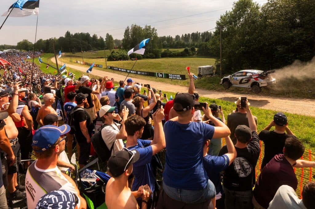 WRC Delfi Rally Estonia katsel sõitev Rally1 auto, Elfyn Evans, möödub suurest publikualast, kus pealtvaatajad lehvitavad Eesti lippe ja jäädvustavad sõitu telefonidega.