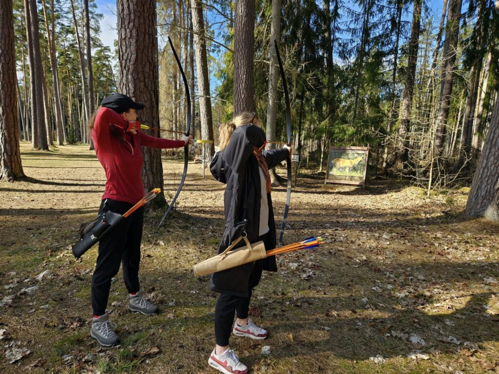 Two women practicing archery in a pine forest